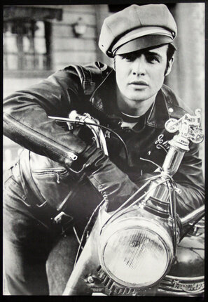 a man in leather jacket and hat leaning on a motorcycle